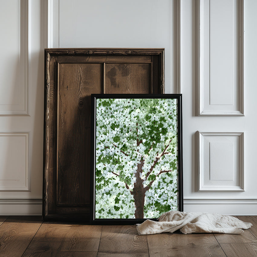 Framed artwork of a tree with white flowers on a wooden floor against a white paneled wall.