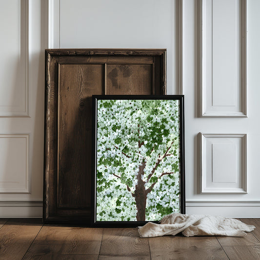 Framed artwork of a tree with white flowers on a wooden floor against a white paneled wall.