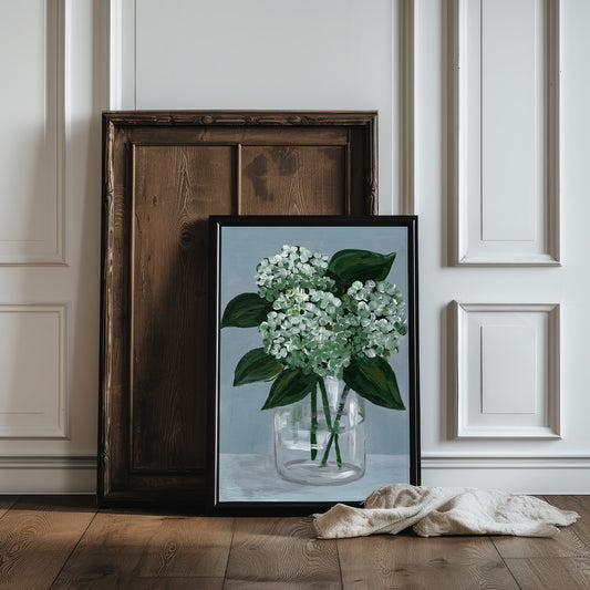 Framed artwork of a floral arrangement on a wooden floor with a white paneled wall background.